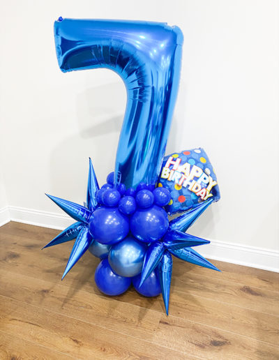 A blue number 7 balloon surrounded by matching blue star-shaped balloons, with a "happy birthday" balloon on a wooden floor, styled by a professional balloon decorator in Nebraska.