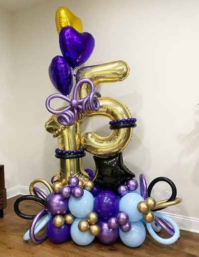 A colorful balloon display crafted by a skilled balloon decorator featuring a large number "8" in gold, surrounded by purple, blue, and golden balloons, some shaped like hearts, arranged against a beige wall
