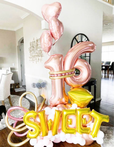 Decorative display featuring large "16" and "sweet" balloons in a home setting, with pink and gold colors, accompanied by a balloon arch and additional heart-shaped balloons.