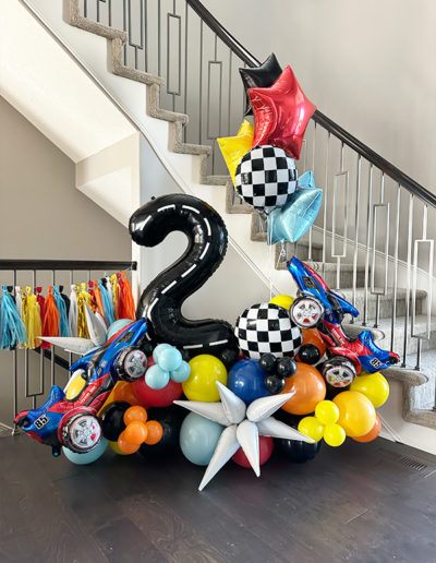 Colorful balloon arch display for a 2nd birthday in Omaha, featuring a large black '2', race cars, stars, and checkered patterns under a staircase.