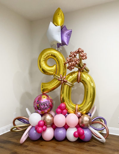 A colorful balloon garland featuring large golden numerals "90" with an assortment of pink, purple, and white balloons and a "happy birthday" balloon.