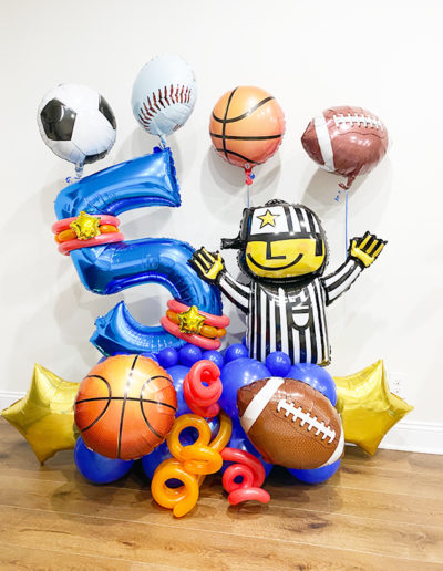 Sports-themed party balloons, featuring a referee, numbers 5 and 2, and various sports balls, assembled into a balloon arch in a room with wooden flooring.
