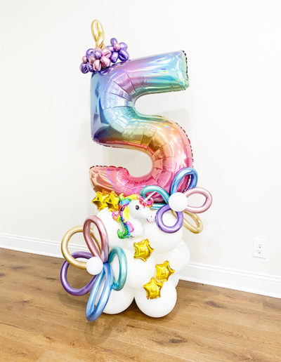 Colorful number "5" balloon display with a unicorn-themed decoration and small gold star balloons on a white cloud base against a plain wall in Omaha.