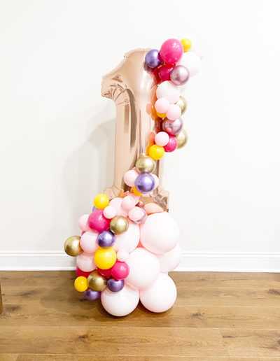 A balloon sculpture shaped like the number one, crafted by a talented balloon decorator in Omaha, Nebraska, featuring a mix of colorful balloons on a wooden floor against a white wall.