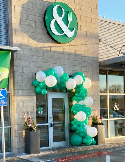 Store entrance adorned with a balloon garland of green and white balloons, highlighted by a green logo with an ampersand (&) mounted on a stone facade.