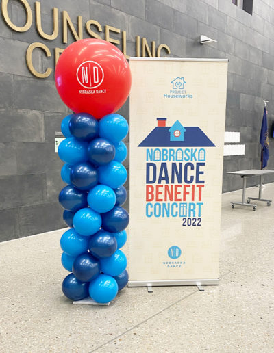 A display promoting the Nebraska dance benefit concert 2022, featuring a tall blue and red balloon arch beside a banner with logos and event details.