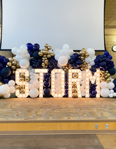 An event display in Omaha with the word "storm" spelled out in large, illuminated letters, surrounded by clusters of white, blue, and gold balloons crafted by a professional balloon decorator.