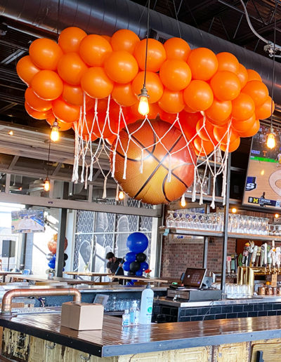 A sports-themed bar decorated with a basketball hoop and orange balloons overhead, designed by a balloon decorator from Nebraska, with a bar counter and stools in the foreground.