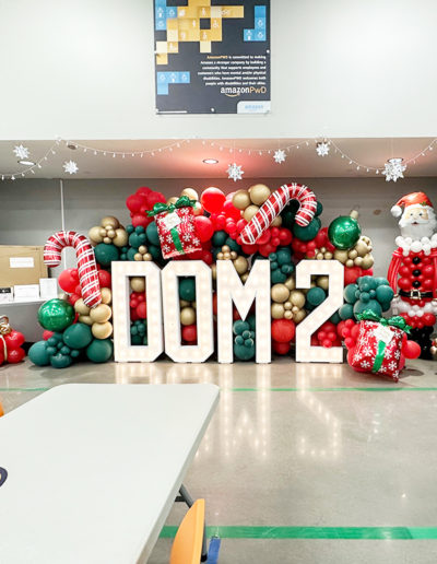 Decorative display at Amazon's office with "dom 2" sign surrounded by festive holiday elements including oversized ornaments, gifts, a Santa figure, and a balloon garland.