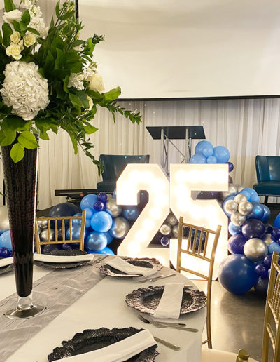 Elegant event setup featuring a large "25" illuminated centerpiece, surrounded by blue and silver balloons, with decorated dining tables and floral arrangements.
