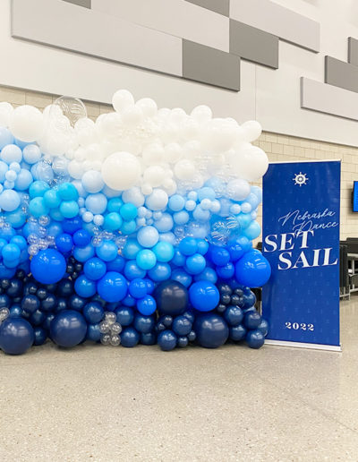 A banner reading "Omaha set sail 2023" next to a large balloon arch in various shades of blue and white, crafted by a professional balloon decorator in an indoor setting.