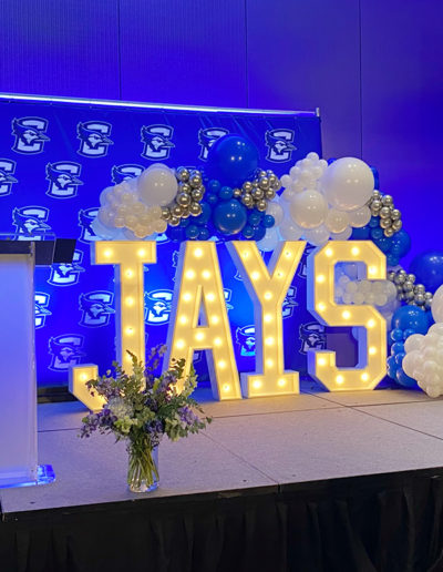 A decorated stage featuring the word "jays" in large illuminated letters, surrounded by a balloon arch of blue and white balloons, at an event with a floral arrangement in front.