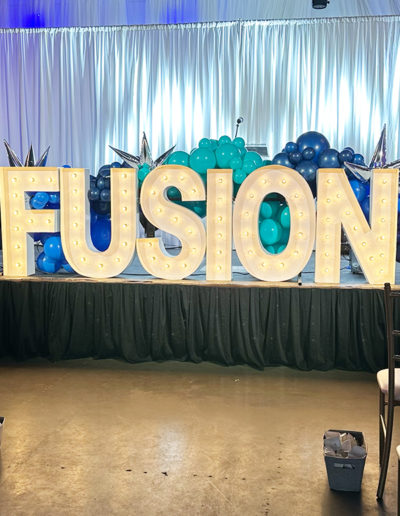 Large illuminated "fusion" sign displayed on stage, flanked by a balloon arch in shades of blue and teal, with curtains in the background.
