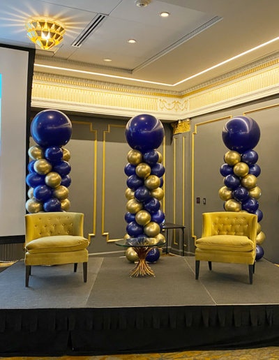 Elegant event room with yellow chairs and tall balloon columns in blue and gold, matching ornate gold-trimmed walls and ceiling.