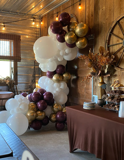 Balloon arch in white, gold, and purple beside a dessert table in a rustic room with wooden walls.