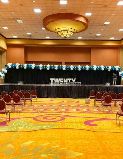 A conference hall is set up for an event with rows of red chairs facing a stage under a banner reading "twenty", decorated with blue and white balloon garland.