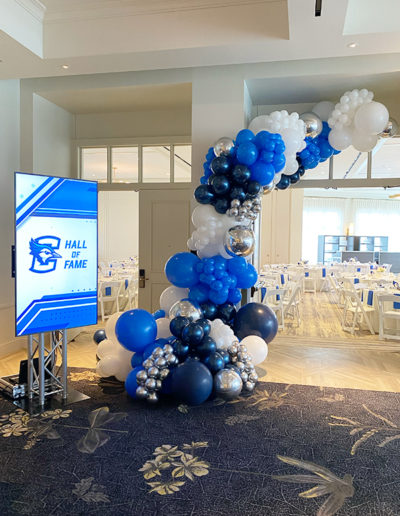 An event hall decorated with a blue and white balloon garland next to a digital display board with a "hall of fame" graphic, surrounded by arranged tables and chairs.