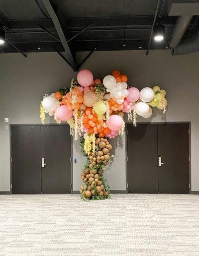 A vibrant balloon arch crafted by a skilled balloon decorator in shades of pink, white, and gold, hovering above black double doors in a room with gray carpet.