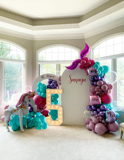 Indoor birthday setup featuring a large balloon arch with a unicorn and "be magical" decor, complemented by light-up letters spelling "sanaya".