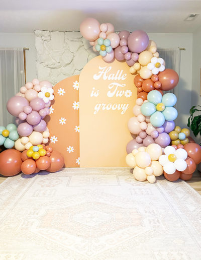 A colorful party setup featuring a balloon garland in pastel shades and a large peach backdrop with the phrase "halle is two groovy" in retro-styled lettering.
