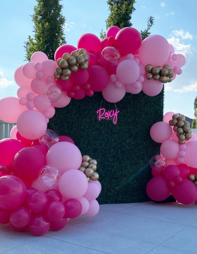 A decorative balloon arch made of pink and gold balloons with the name "Roxy" in cursive, set against a backdrop of a green hedge in Omaha, Nebraska.