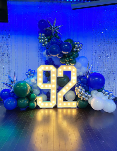 A festive display featuring the number 92 in large, illuminated numerals, surrounded by clusters of blue and silver balloons and a balloon arch, with star decorations against a blue curtain backdrop with string lights.