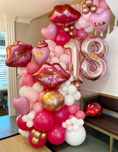 A vibrant balloon arrangement by a balloon decorator for a birthday in Nebraska, featuring red hearts, pink balloons, and gold '18' balloons next to a staircase.