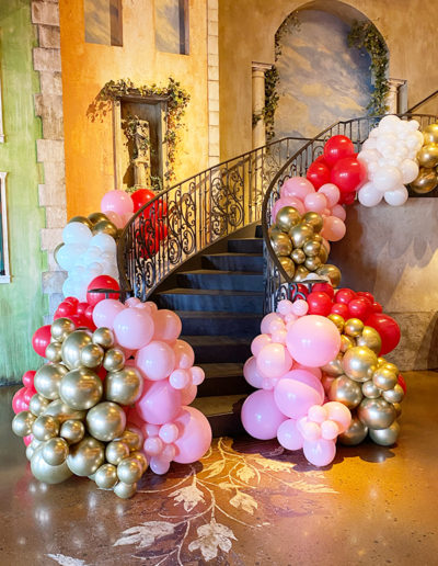An ornate staircase adorned with a balloon arch featuring clusters of pink, red, and gold balloons in an elegantly designed room.