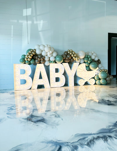 Decorative display featuring the lit-up word "baby" in large letters, surrounded by a balloon garland in white, gold, and blue colors.