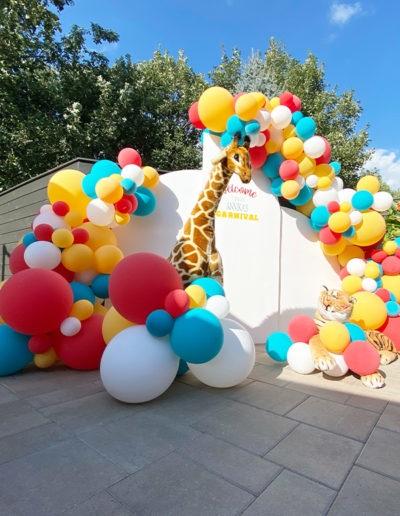 A colorful carnival display features a tall giraffe model peeking through an arch of multicolored balloons, enhanced by a balloon garland, and a tiger model lying nearby.