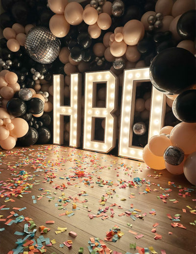 Large "hbo" light-up letters surrounded by a balloon garland and colorful confetti on a wooden floor, crafted by a skilled balloon decorator.