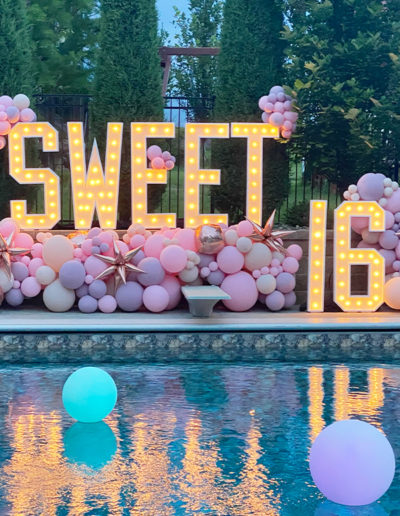 Sweet 16" lit sign adorned with a balloon garland, featuring pink and white balloons by a pool at twilight, reflecting colorful lights on the water.