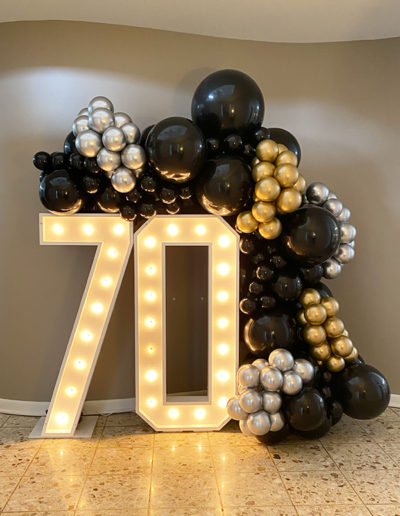 A large decorative numeral "70" with integrated lights, surrounded by a balloon arch comprised of black, silver, and gold balloons.