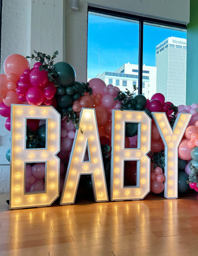 Large lighted letters spelling "baby" in front of a colorful balloon arch with Omaha's cityscape visible through the window.