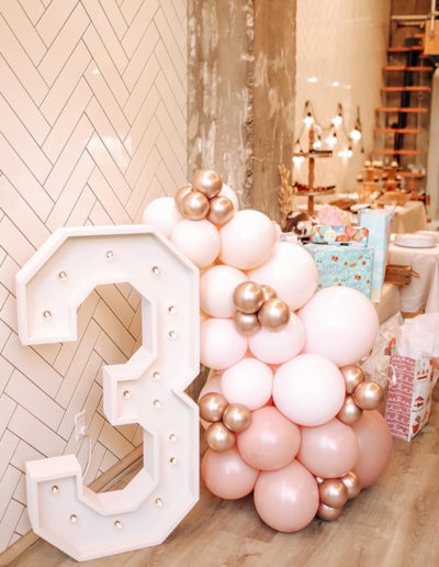 A large, white number "3" adorned with balloon garlands and decorated by a balloon decorator, positioned in a boutique shop.