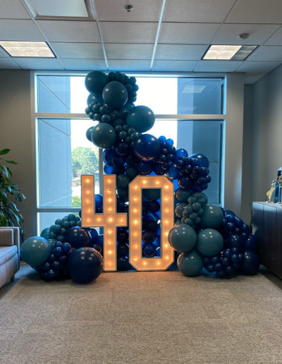 Large "40" illuminated by lights surrounded by clusters of blue balloons and a balloon arch in an office setting with a window in the background.