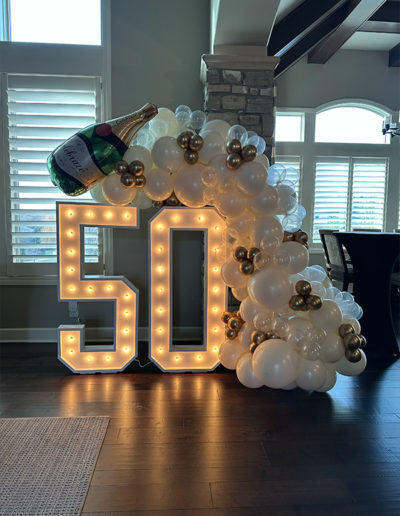 Large decorative light-up numbers "50" adorned with a balloon arch featuring gold and white balloons and a champagne bottle, set in a home interior, designed by a skilled balloon decorator.
