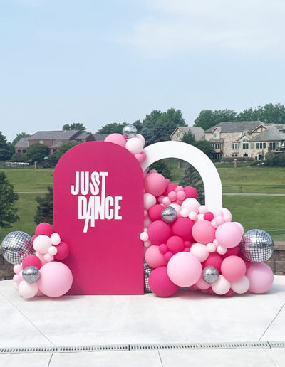 Outdoor display featuring a large pink sign reading "just dance" surrounded by various pink and silver balloons and two large white arcs, created by a balloon decorator, set against a residential backdrop.