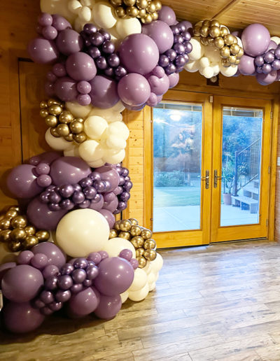 Wooden interior space with double doors framed by a balloon arch made of purple and gold balloons.